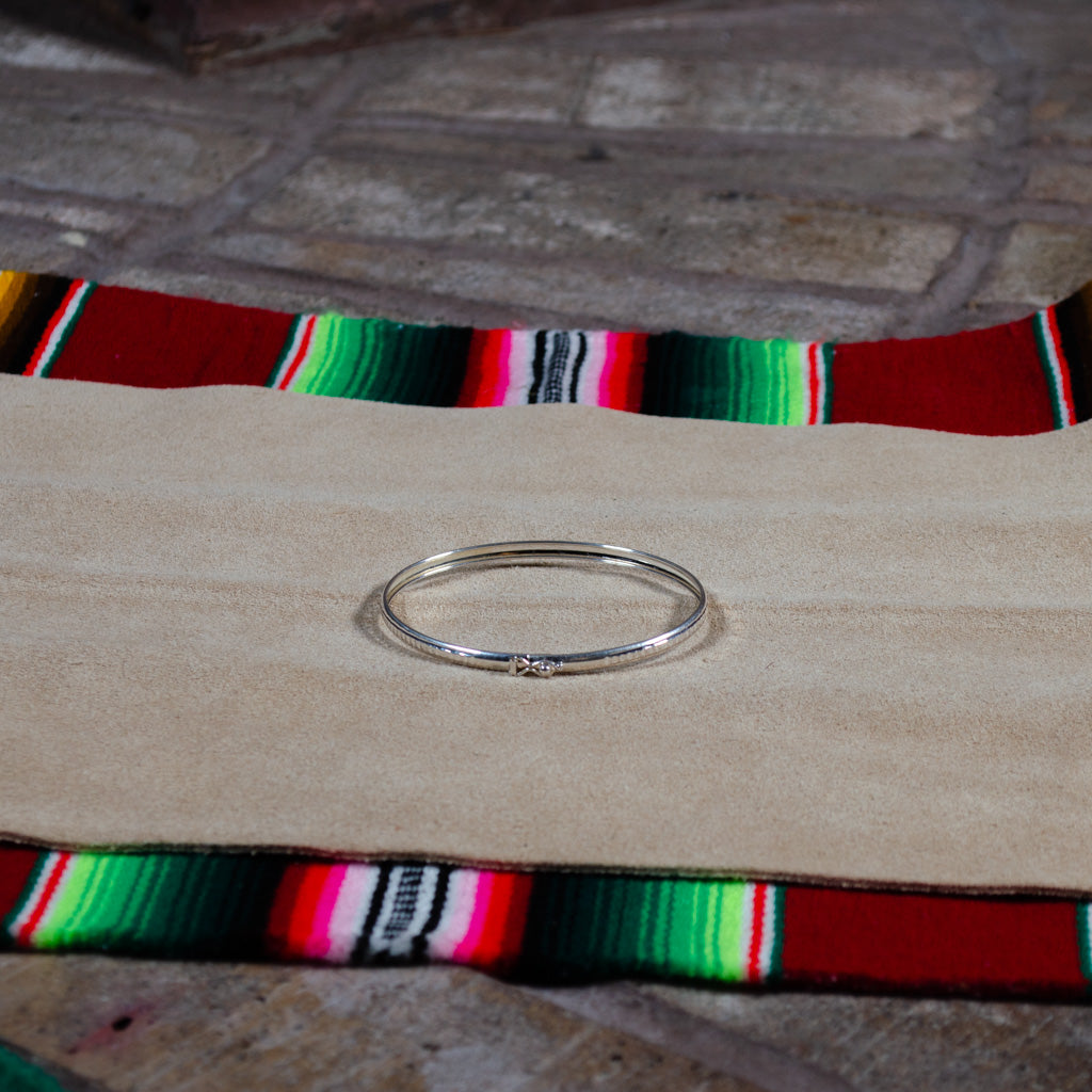 A thin silver bangle bracelet with a tiny central stone, resting on a beige suede-like pad over a colorful red, green, and black patterned textile.